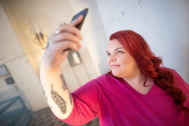 Satu ottamassa selfiekuvaa itsestään. Hänellä on kirkkaan punaiset hiukset ja fuksian punainen paita.
