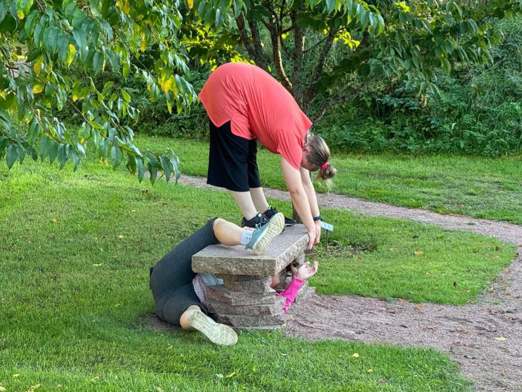 Parkour-tädit puiston penkillä harrastamassa. Yksi venyttelee punaisessa paidassa kaksin kerroin seisoen penkillä, toinen on ryömimässä penkin alta. Ympärillä kaupunkipuisto kesällä.